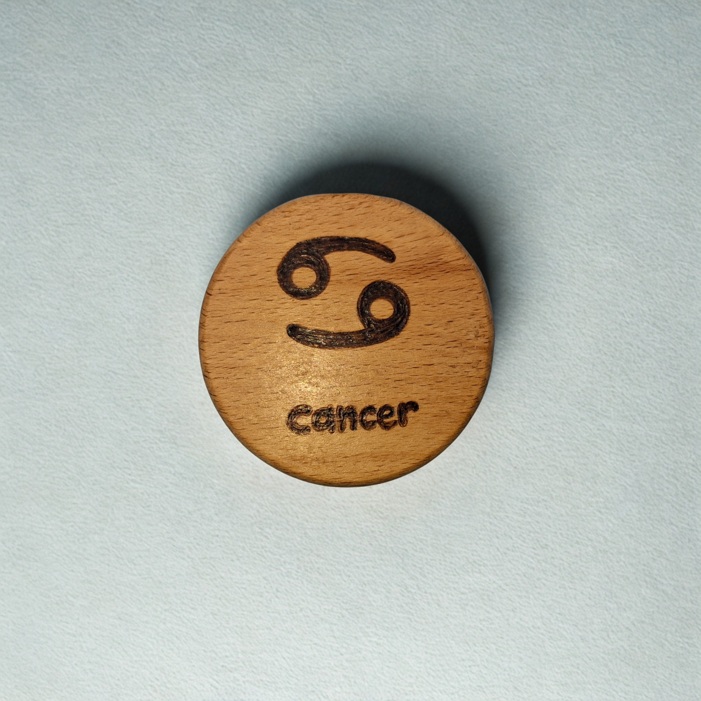 Close-up of a light-colored beechwood circle, 5cm in diameter and 0.4cm thick, featuring the words "Cancer" and an illustration of the star sign symbol, hand-burned into its surface using pyrography. The wood has a smooth, waxed finish and the text is clearly legible.