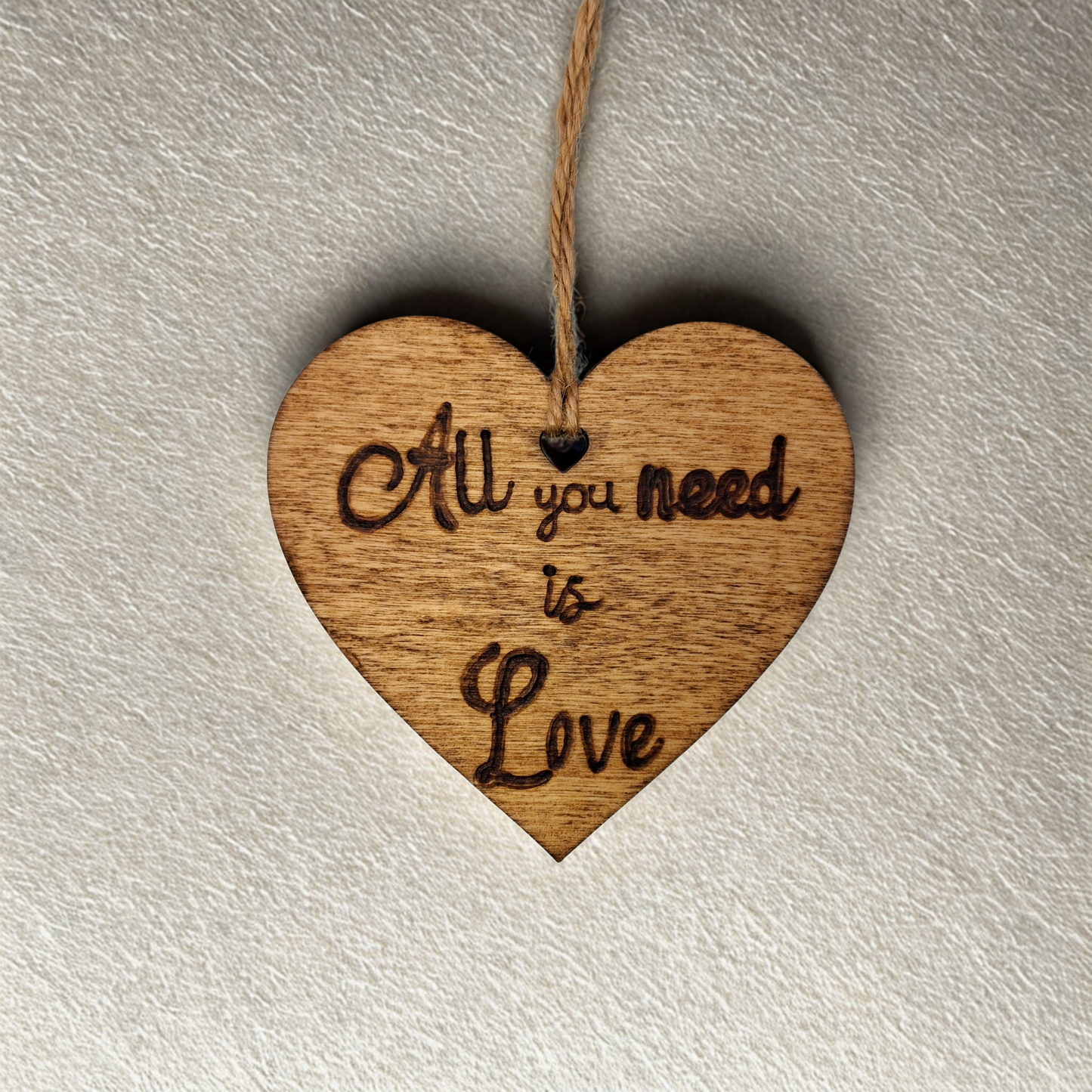 Heart-shaped wooden ornament, hand-drawn with pyrography, featuring the engraved phrase "All you need is Love" in a decorative script, set against a textured background.
