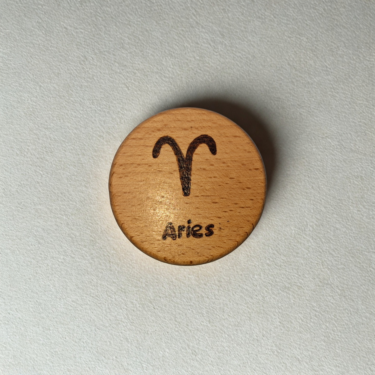 Close-up of a light-colored beechwood circle, 5cm in diameter and 0.4cm thick, featuring the words "Aries" and an illustration of the star sign symbol, hand-burned into its surface using pyrography. The wood has a smooth, waxed finish and the text is clearly legible.