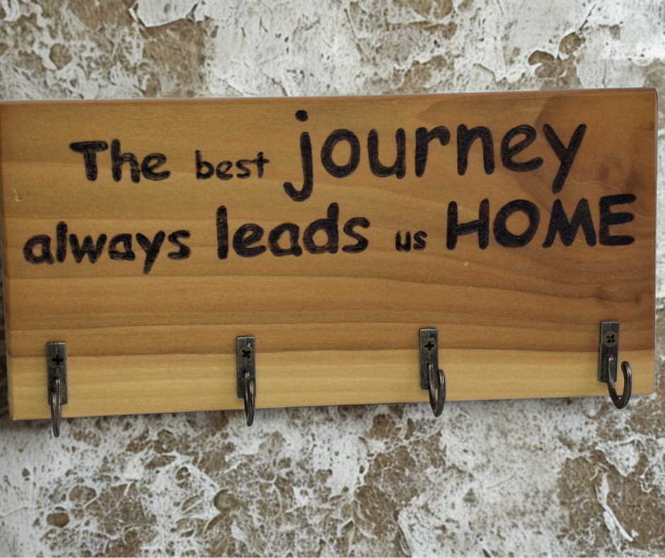 A four-key hook hanger crafted from two-tone Tulipwood, featuring hand-burnt lettering that reads "The best journey always leads us home." The wood has visible grain, showcasing its natural variations.