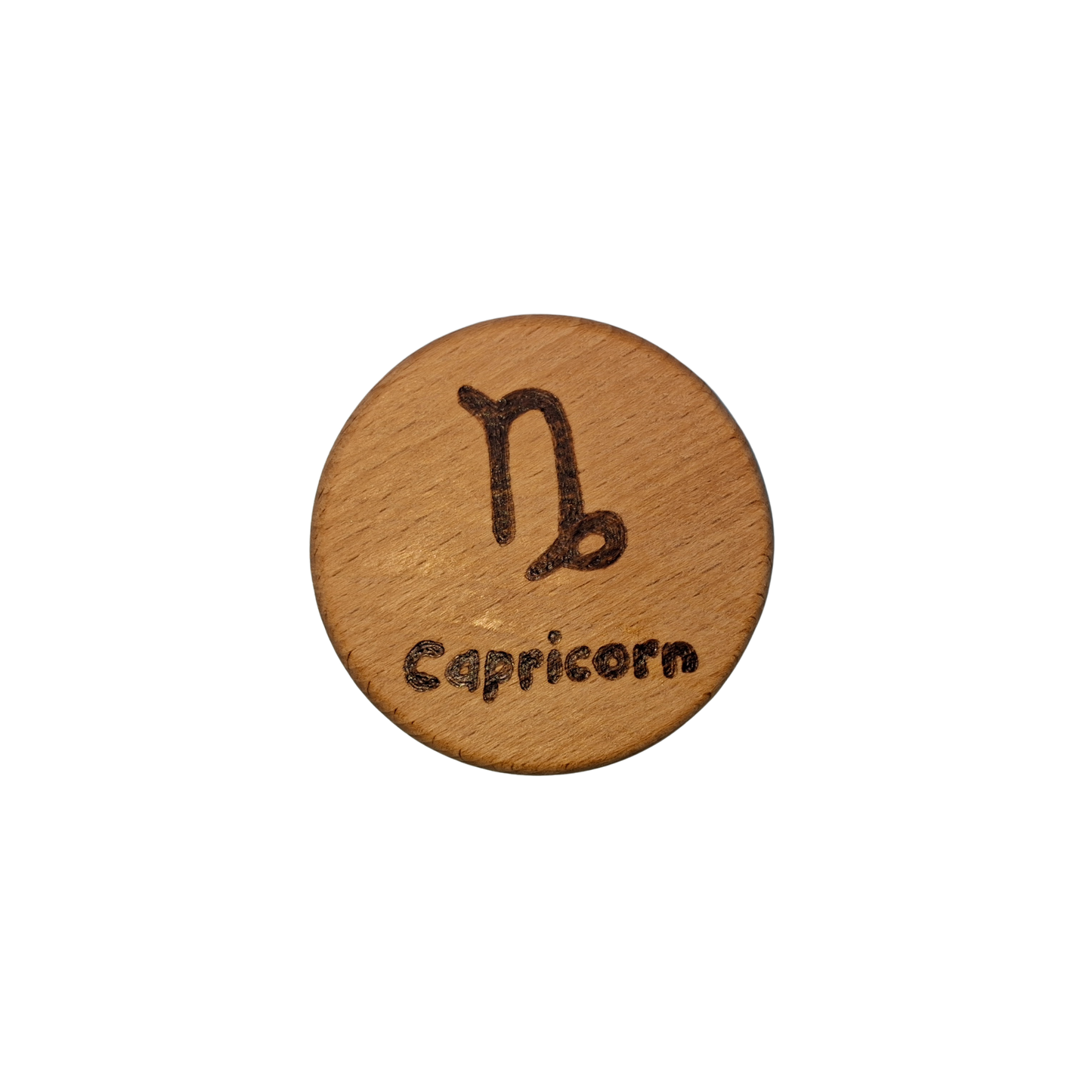 Close-up of a light-colored beechwood circle, 5cm in diameter and 0.4cm thick, featuring the words "Capricorn" and an illustration of the star sign symbol, hand-burned into its surface using pyrography. The wood has a smooth, waxed finish and the text is clearly legible.