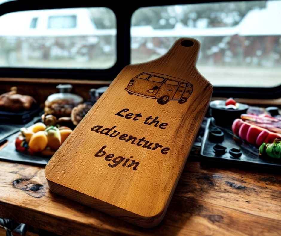 A small, hand-cut beechwood chopping board with hand-decorated pyrography wording "Let the adventure begin" and an illustration of a traditional campervan. It's finished with food-safe hardwax oil.