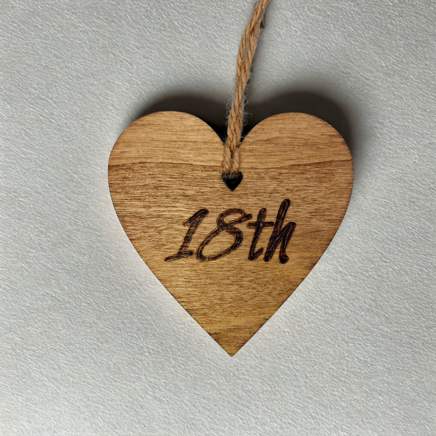A small, 8cm by 8cm, light-colored wooden heart, decorated with "18th" hand-burned into its surface using pyrography. A short length of natural jute rope is tied through the top for hanging.