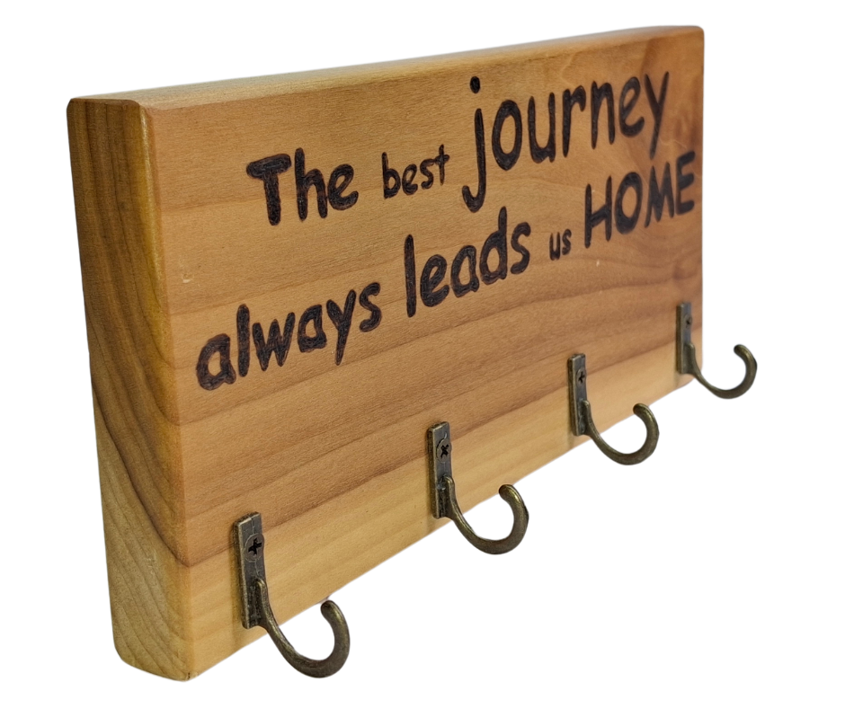 A four-key hook hanger crafted from two-tone Tulipwood, featuring hand-burnt lettering that reads "The best journey always leads us home." The wood has visible grain, showcasing its natural variations.