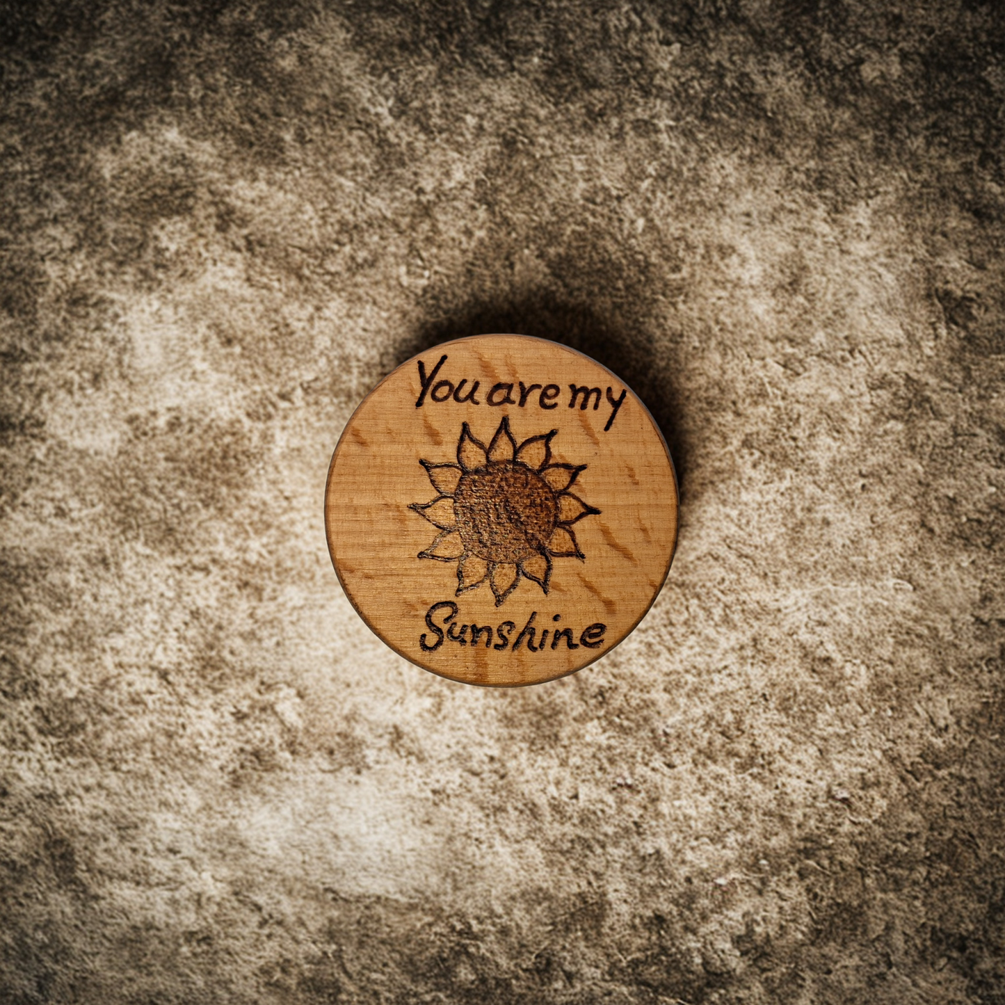 Close-up of a light-colored beechwood circle, 5cm in diameter and 0.4cm thick, featuring the words "You are my sunshine" and an illustration of a sunflower hand-burned into its surface using pyrography. The wood has a smooth, waxed finish and the text is clearly legible.