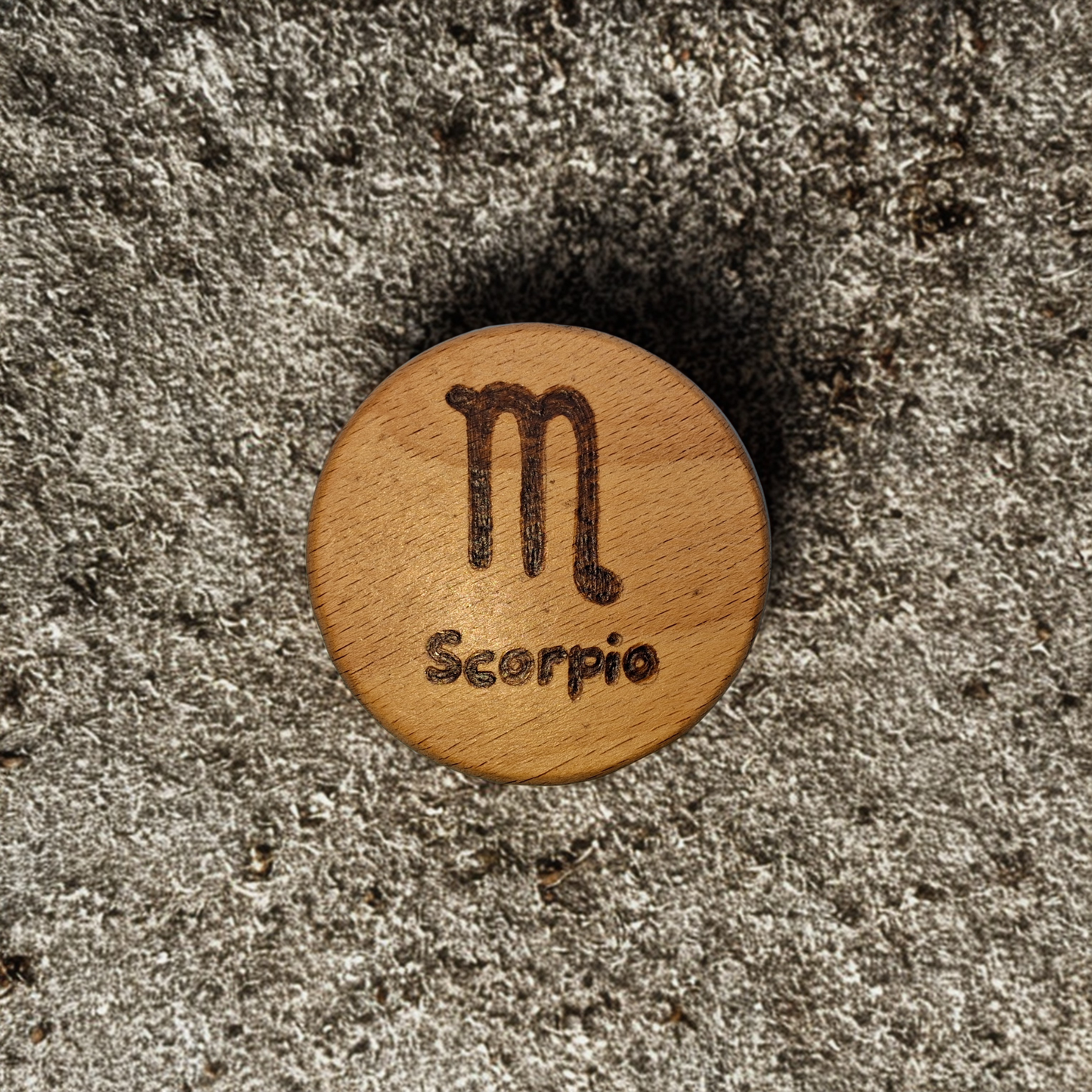 Close-up of a light-colored beechwood circle, 5cm in diameter and 0.4cm thick, featuring the words "Scorpio" and an illustration of the star sign symbol, hand-burned into its surface using pyrography. The wood has a smooth, waxed finish and the text is clearly legible.