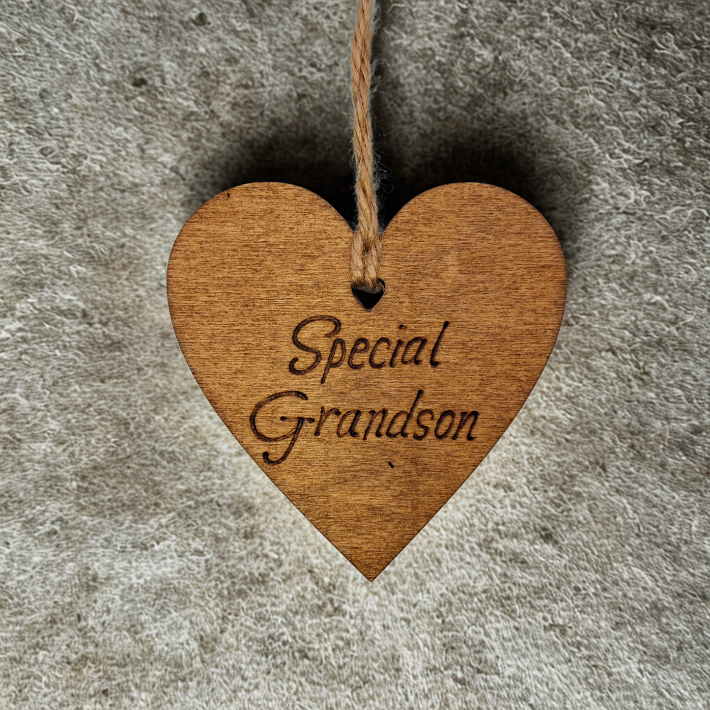 A small, possibly 8cm by 8cm, light-colored wooden heart, decorated with "Special Grandson" hand-burned into its surface using pyrography. A short length of natural jute rope is tied through the top for hanging.