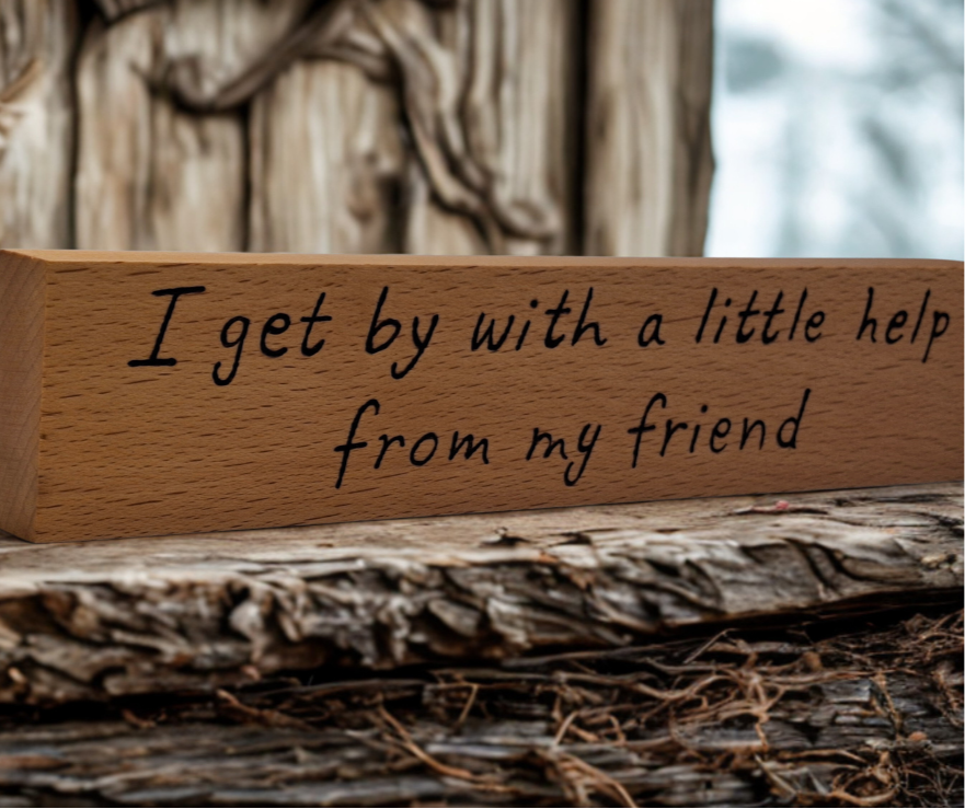 A hand-burnt, freestanding wooden plaque with the words "I get by with a little help from my friend" in a casual, handwritten style. The wood has visible grain and a rustic, natural finish.