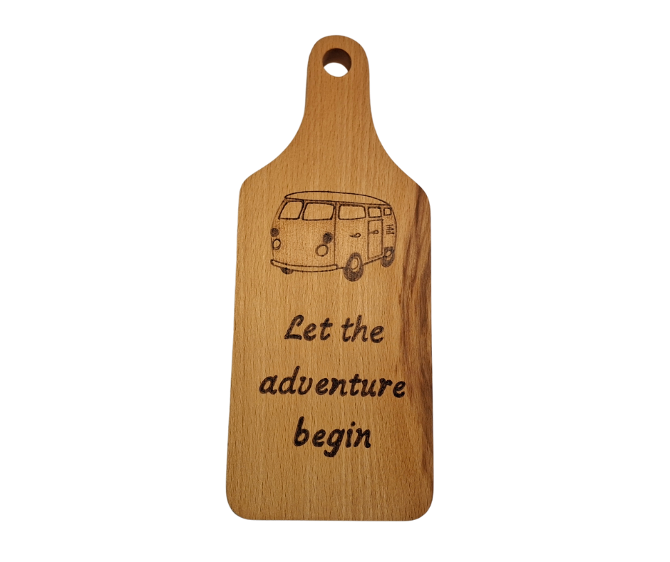 A small, hand-cut beechwood chopping board with hand-decorated pyrography wording "Let the adventure begin" and an illustration of a traditional campervan. It's finished with food-safe hardwax oil.