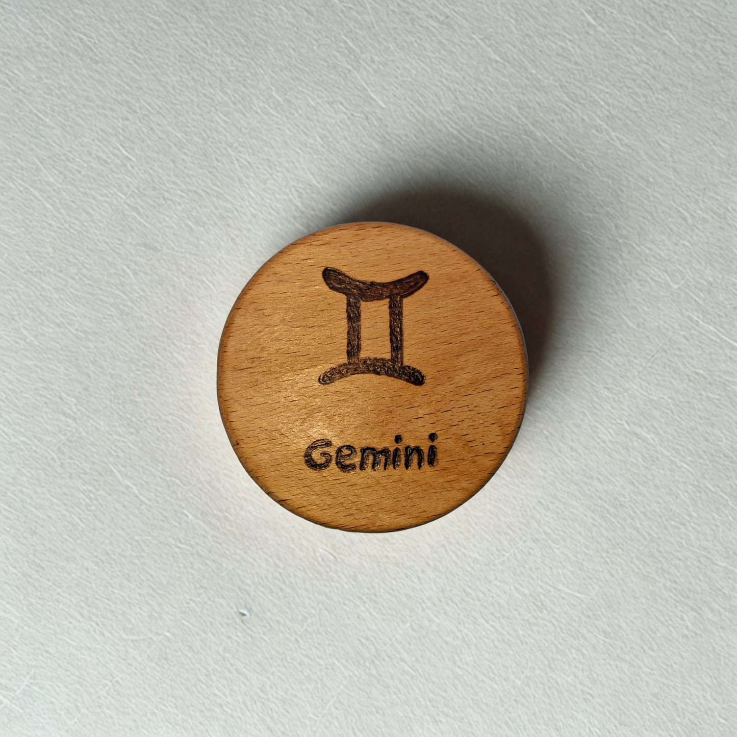 Close-up of a light-colored beechwood circle, 5cm in diameter and 0.4cm thick, featuring the words "Gemini and an illustration of the star sign symbol, hand-burned into its surface using pyrography. The wood has a smooth, waxed finish and the text is clearly legible.