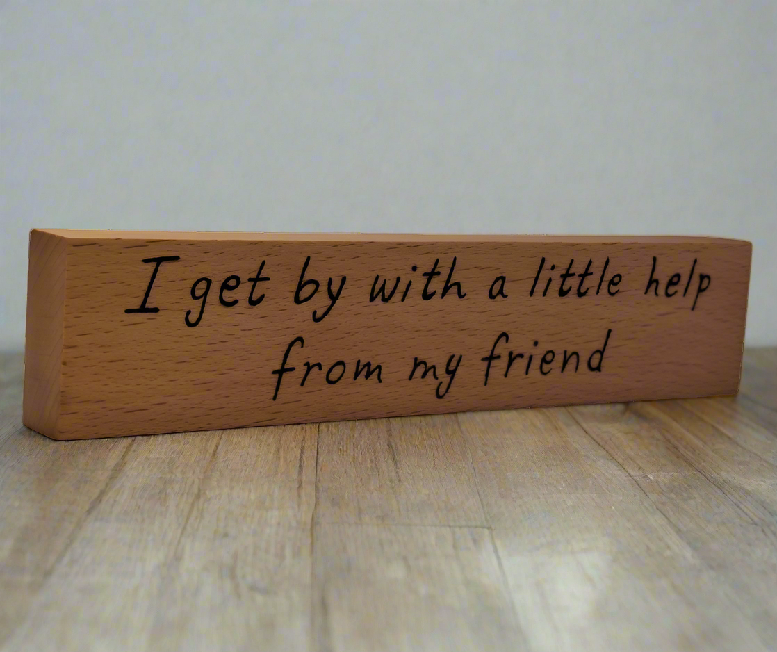 A hand-burnt, freestanding wooden plaque with the words "I get by with a little help from my friend" in a casual, handwritten style. The wood has visible grain and a rustic, natural finish.