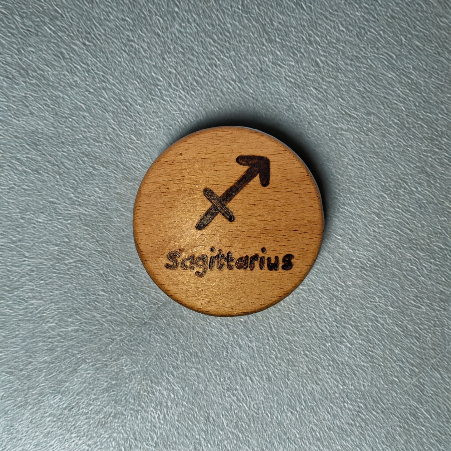 Close-up of a light-colored beechwood circle, 5cm in diameter and 0.4cm thick, featuring the words "Sagittarius" and an illustration of the star sign symbol, hand-burned into its surface using pyrography. The wood has a smooth, waxed finish and the text is clearly legible.