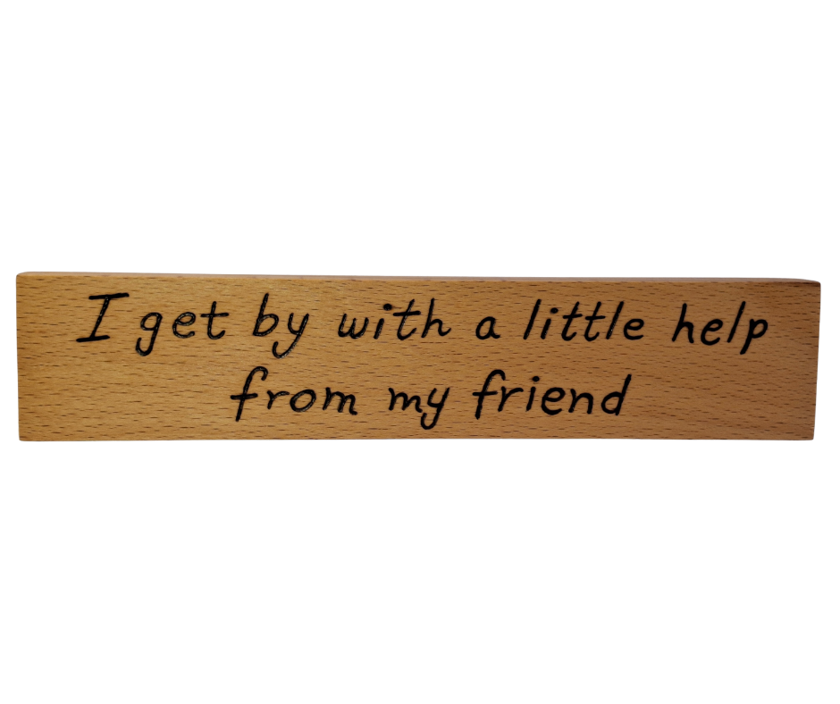 A hand-burnt, freestanding wooden plaque with the words "I get by with a little help from my friend" in a casual, handwritten style. The wood has visible grain and a rustic, natural finish.