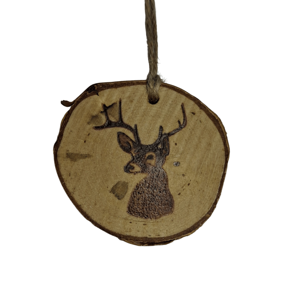 A small Christmas ornament, a wooden log slice with a jute rope for hanging, is centered against a black background. The log slice is decorated with pyrography, featuring a bust of a deer with antlers. The deer's body is filled in with a dark, textured pattern.