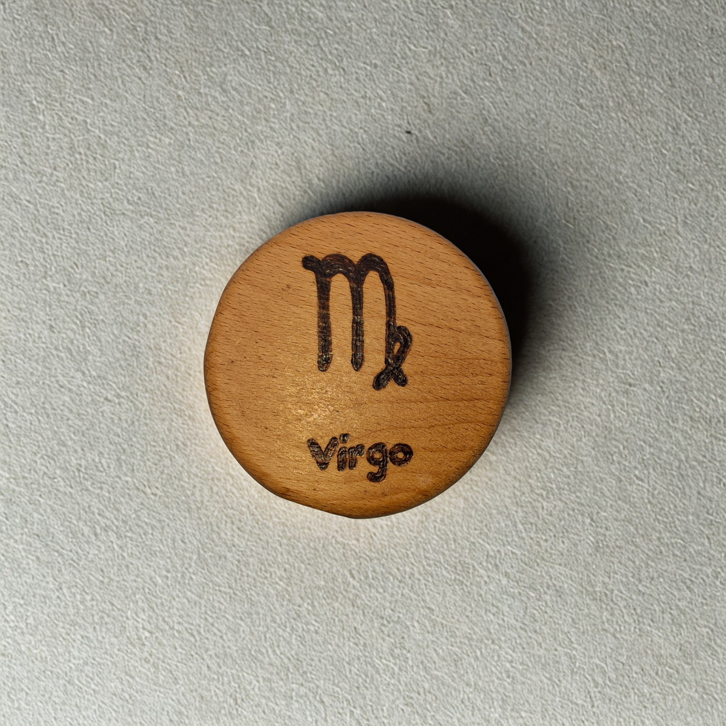 Close-up of a light-colored beechwood circle, 5cm in diameter and 0.4cm thick, featuring the words "Virgo" and an illustration of the star sign symbol, hand-burned into its surface using pyrography. The wood has a smooth, waxed finish and the text is clearly legible.