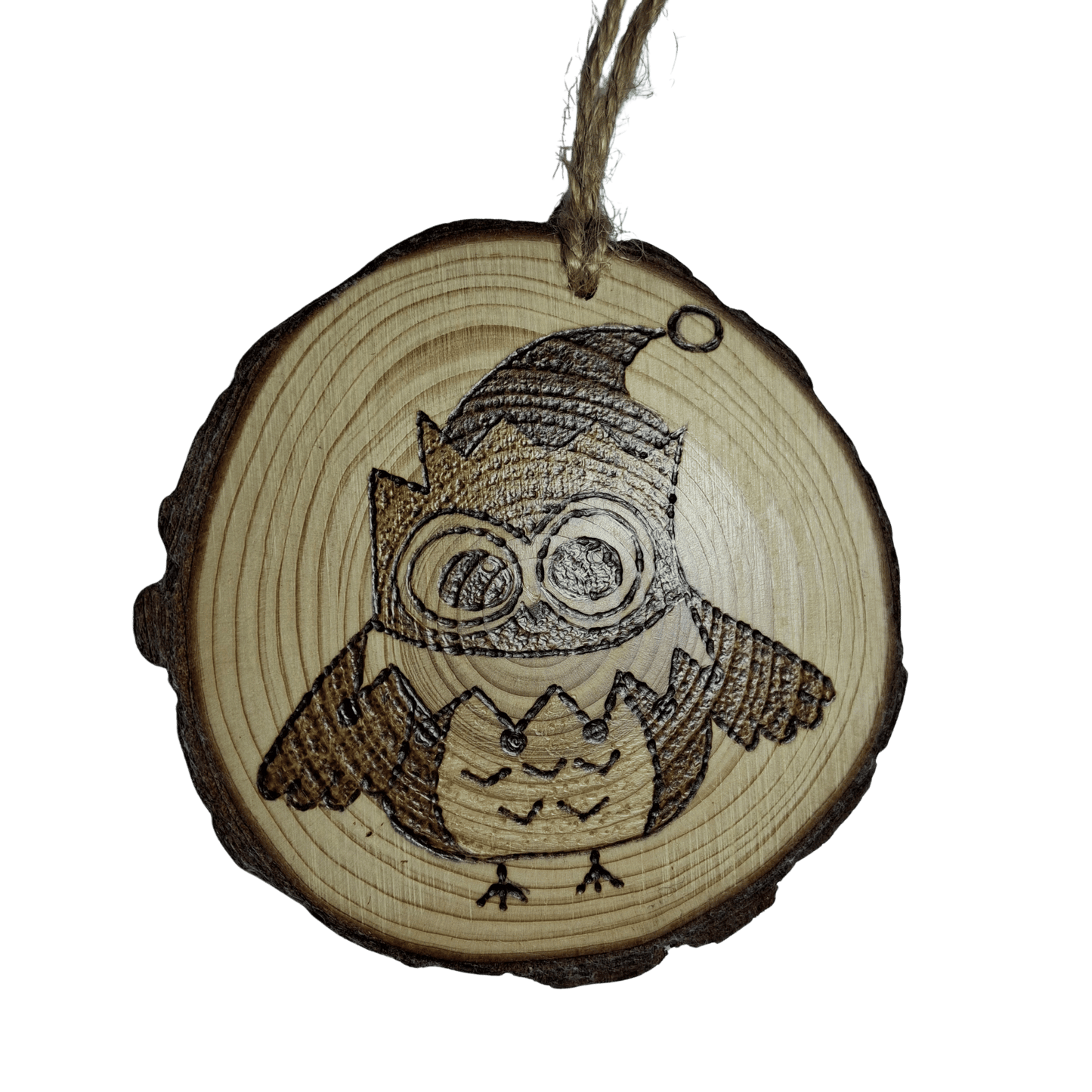 A close-up of a Christmas ornament on a black background. The ornament is a round wooden log slice with a jute rope for hanging. The front of the ornament is decorated with a pyrography design of a cute owl with large eyes, wearing a pointed hat.