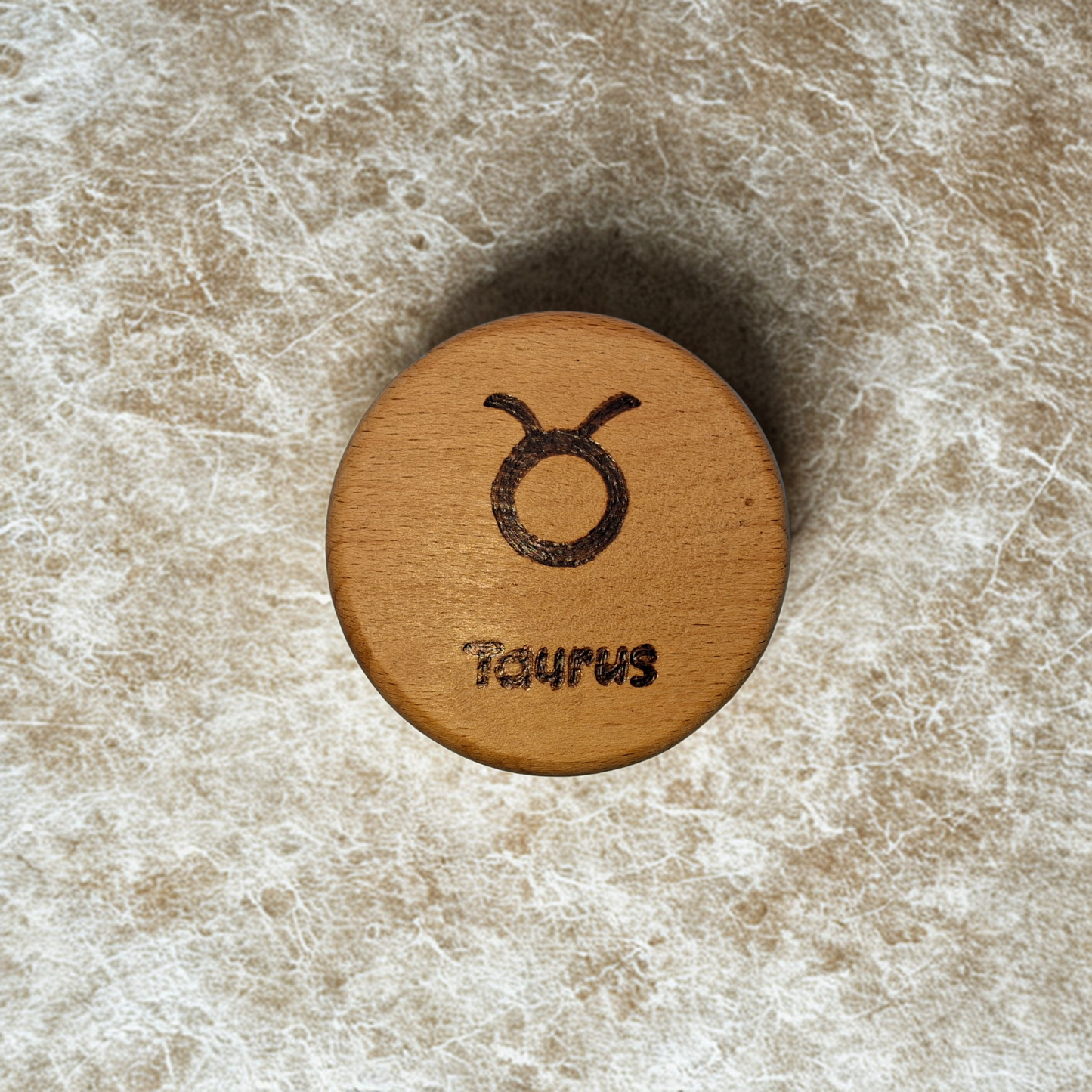 Close-up of a light-colored beechwood circle, 5cm in diameter and 0.4cm thick, featuring the words "Taurus" and an illustration of the star sign symbol, hand-burned into its surface using pyrography. The wood has a smooth, waxed finish and the text is clearly legible.