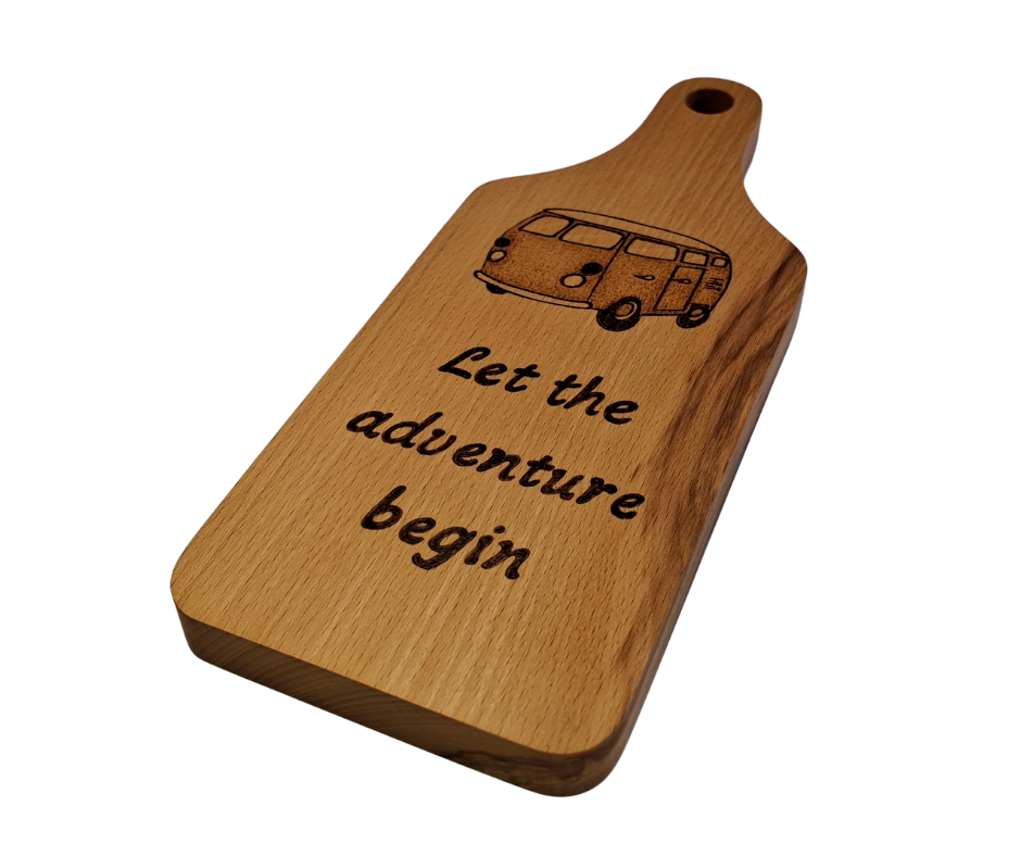 A small, hand-cut beechwood chopping board with hand-decorated pyrography wording "Let the adventure begin" and an illustration of a traditional campervan. It's finished with food-safe hardwax oil.