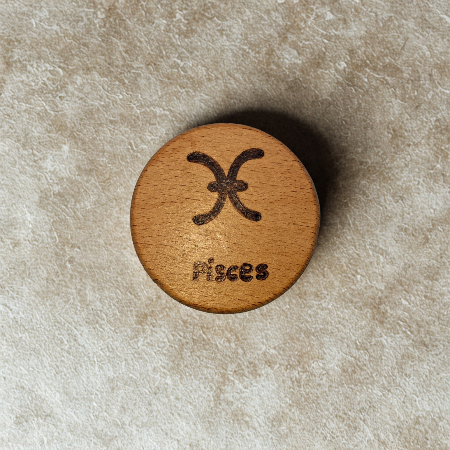 Close-up of a light-colored beechwood circle, 5cm in diameter and 0.4cm thick, featuring the words "Pisces" and an illustration of the star sign symbol, hand-burned into its surface using pyrography. The wood has a smooth, waxed finish and the text is clearly legible.