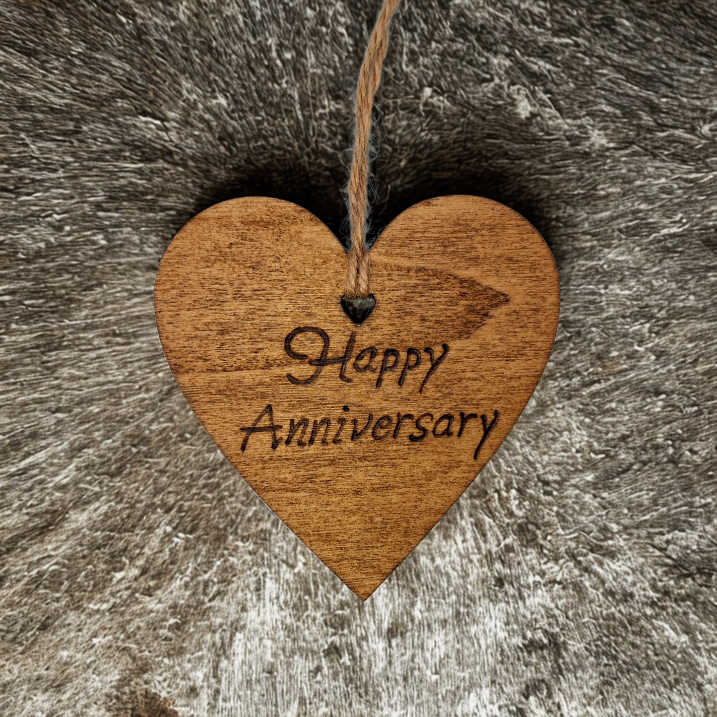 Heart-shaped wooden ornament, hand-drawn with pyrography, featuring the engraved phrase "Happy Anniversary" in a decorative script, set against a textured background.