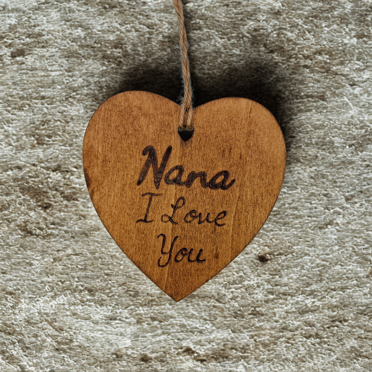 Heart-shaped wooden ornament, hand-drawn with pyrography, featuring the engraved phrase "Nana I Love you" in a decorative script, set against a textured background.