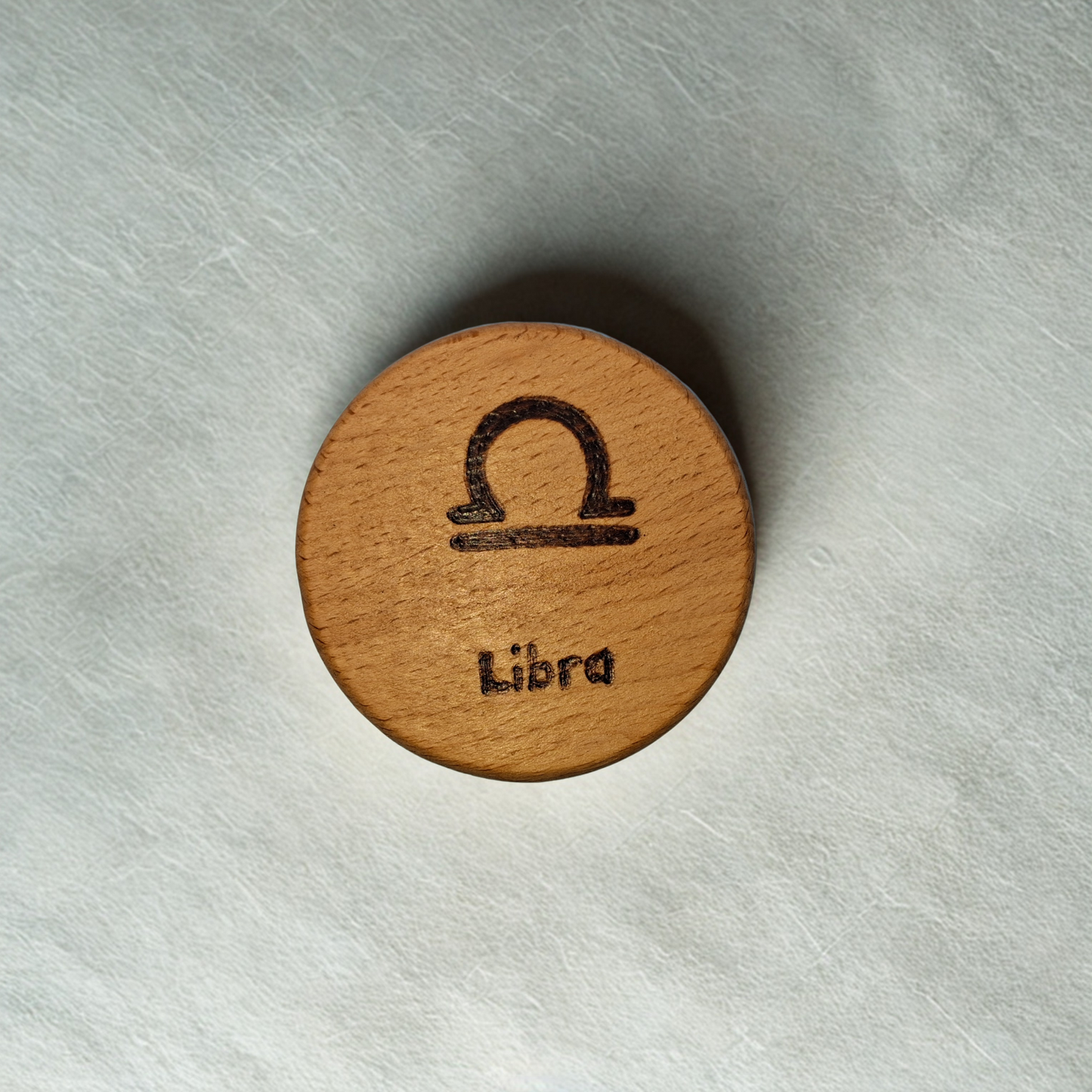 Close-up of a light-colored beechwood circle, 5cm in diameter and 0.4cm thick, featuring the words "Libra" and an illustration of the star sign symbol, hand-burned into its surface using pyrography. The wood has a smooth, waxed finish and the text is clearly legible.