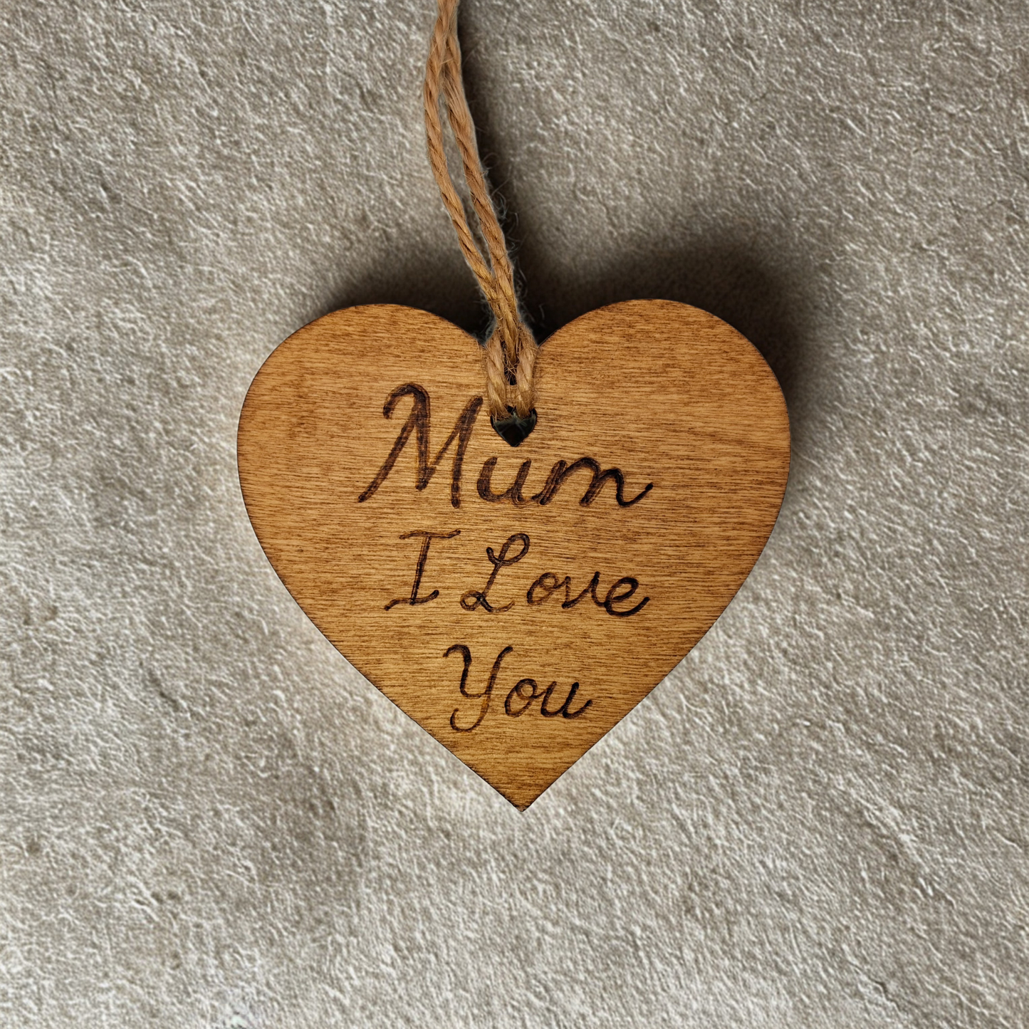 Heart-shaped wooden ornament, hand-drawn with pyrography, featuring the engraved phrase "Mum I love you" in a decorative script, set against a textured background.