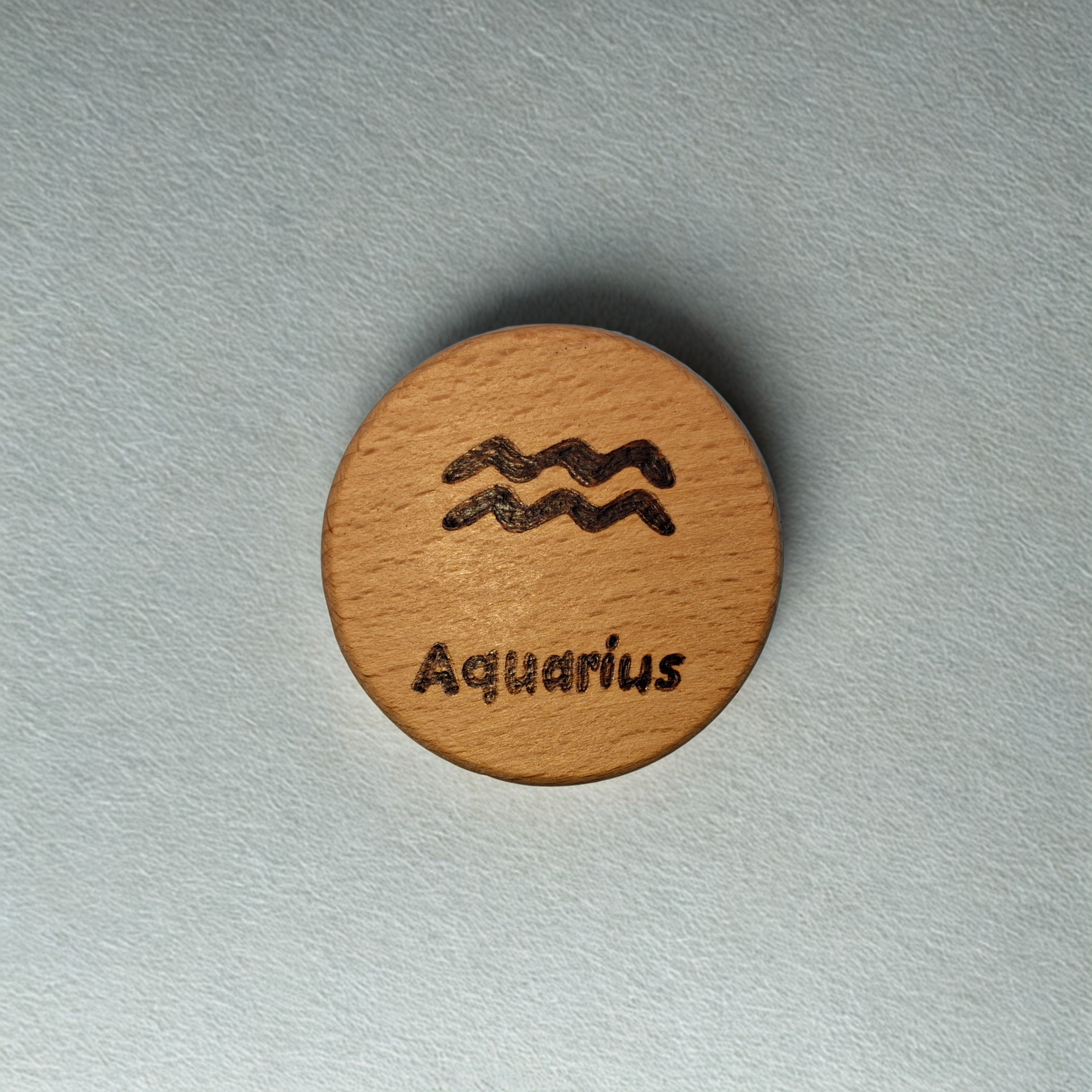 Close-up of a light-colored beechwood circle, 5cm in diameter and 0.4cm thick, featuring the words "Aquarius" and an illustration of the star sign symbol, hand-burned into its surface using pyrography. The wood has a smooth, waxed finish and the text is clearly legible.