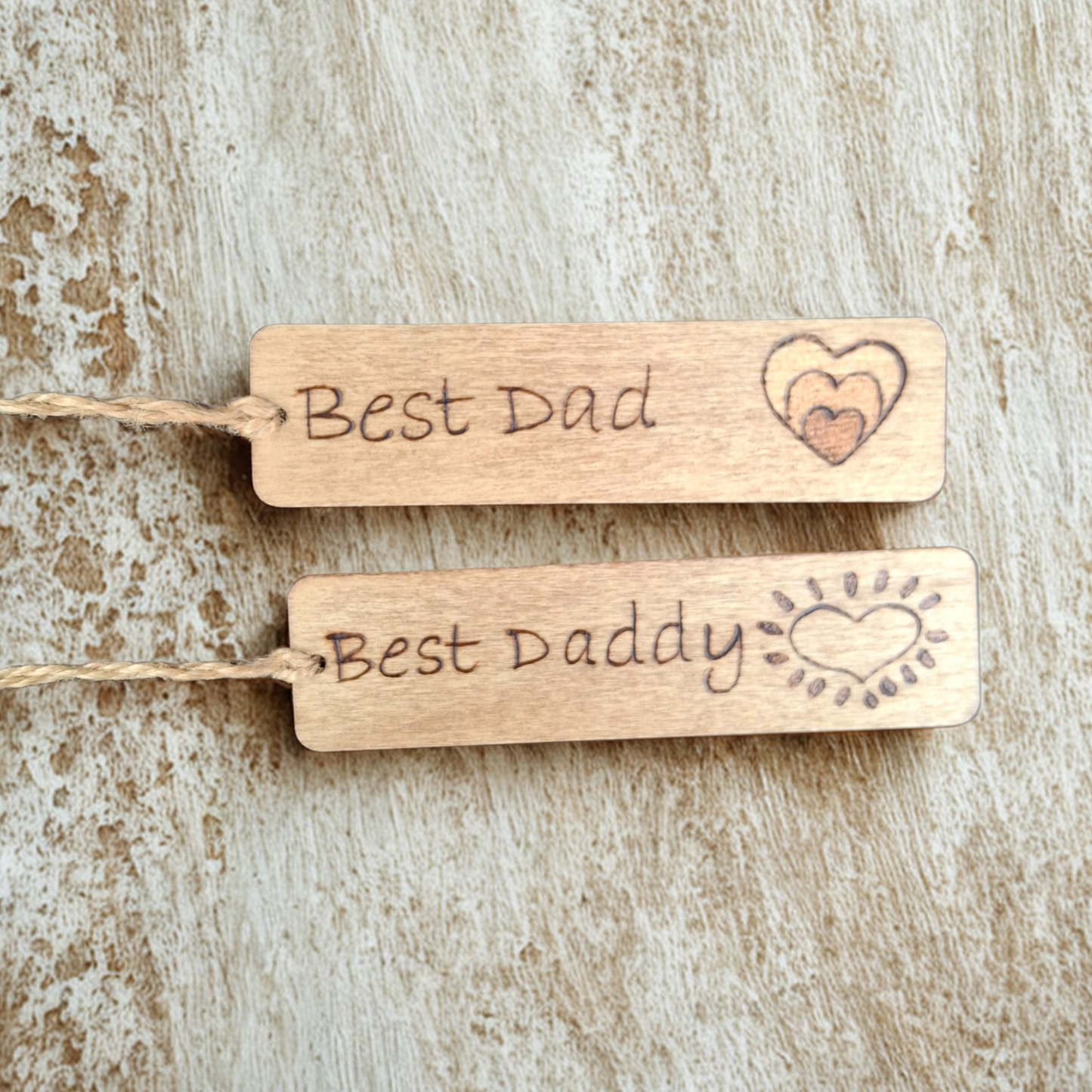 Two wooden bookmarks with string, lying on a light-colored, textured surface. The top tag is engraved with "Best Dad" and three nested hearts. The bottom tag is engraved with "Best Daddy" and a heart surrounded by radiating lines, resembling a sun.