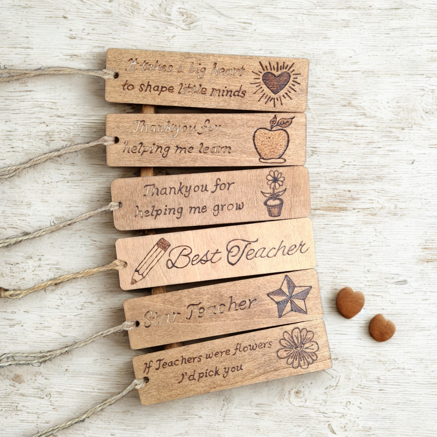 Six rectangular wooden pyrography bookmarks, each with a twine loop on the left, are fanned out diagonally on a light-colored, textured wooden surface. To the right of the bottom bookmark are two small heart-shaped wooden beads. Each bookmark features burned-in text and a design:
From top to bottom:
* Text: "It takes a big heart to shape little minds", Design: A heart with radiating lines (like a sun).
* Text: "Thank you for helping me learn", Design: An apple.
* Text: "Thank you for helping me grow", De