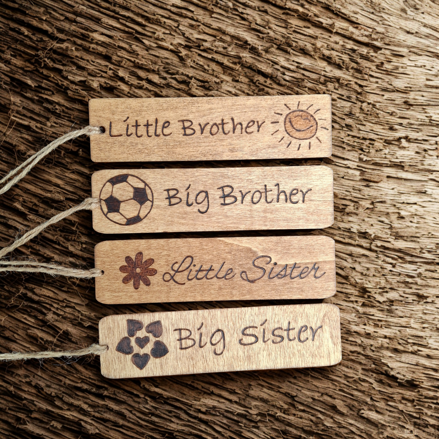 Four wooden bookmarks with twine, lying on a dark, textured wooden surface. From top to bottom, the tags are engraved with: 1. "Little Brother" and a smiling sun. 2. "Big Brother" and a football. 3. "Little Sister" and a flower. 4. "Big Sister" and a cluster of four hearts.