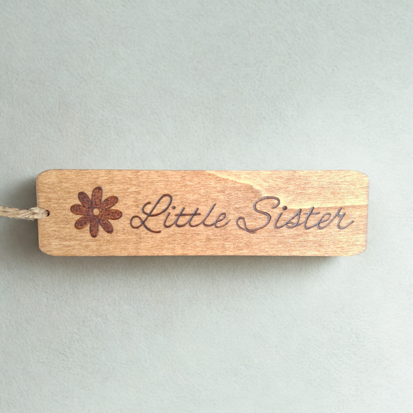 A close-up of a single wooden bookmark with a small piece of twine attached to its left side, lying on a plain, light grey surface. The tag is engraved with a flower design on the left and the words "Little Sister" in a cursive font on the right.
