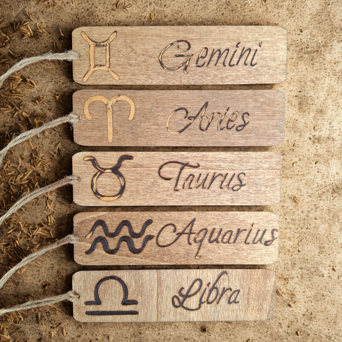 Five wooden bookmarks, each with a twine loop on the left, are arranged vertically on a textured, light brown surface that appears to have some dried plant matter scattered on it. Each tag features a burned-in zodiac symbol on the left and the corresponding zodiac name in a cursive, burned-in font on the right. From top to bottom, the tags display:
* Gemini symbol and "Gemini"
* Aries symbol and "Aries"
* Taurus symbol and "Taurus"
* Aquarius symbol and "Aquarius"
* Libra symbol and "Libra"