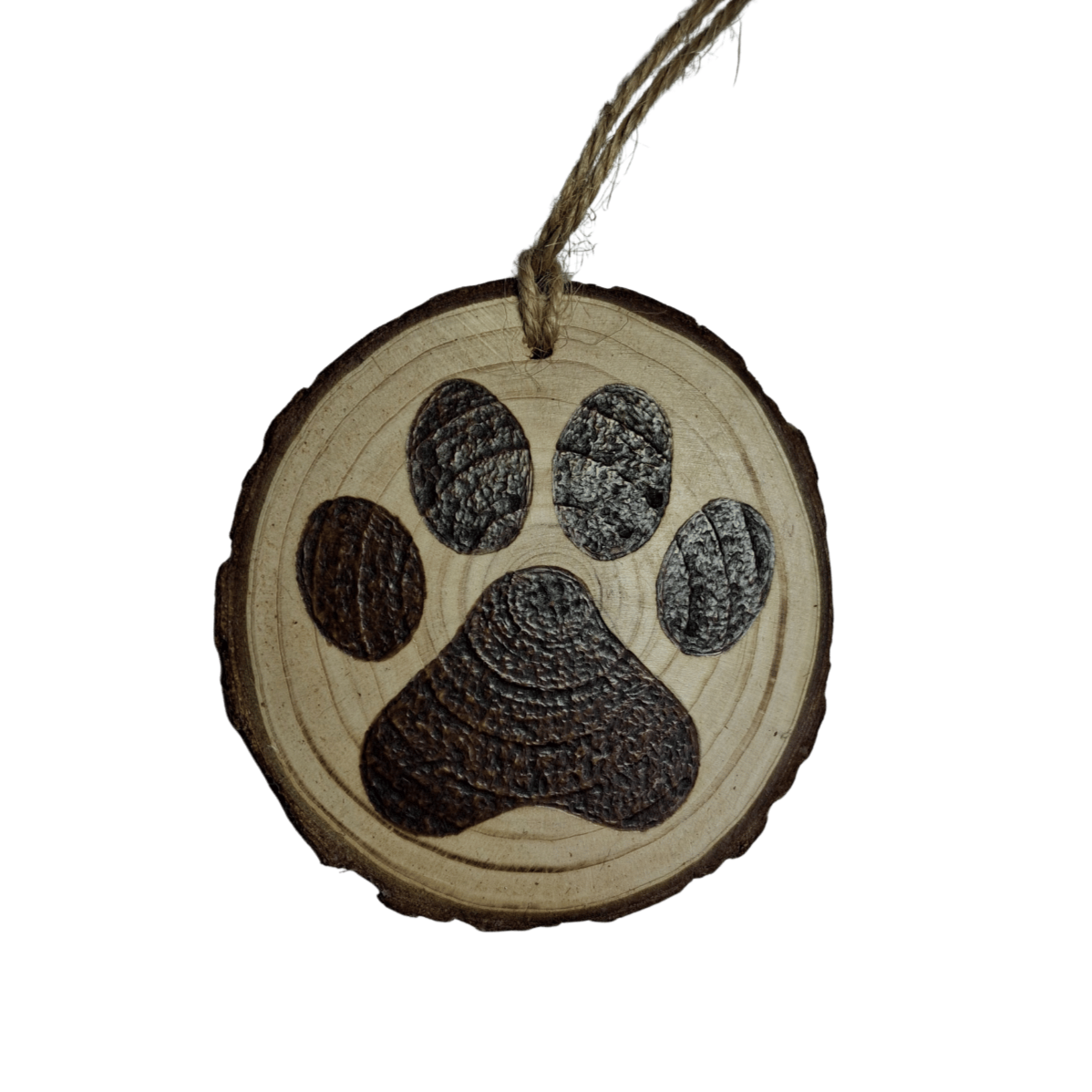 A Christmas ornament, a small, round wooden log slice with a jute rope for hanging, is centered against a black background. The ornament is decorated with a pyrography design of a detailed paw print.