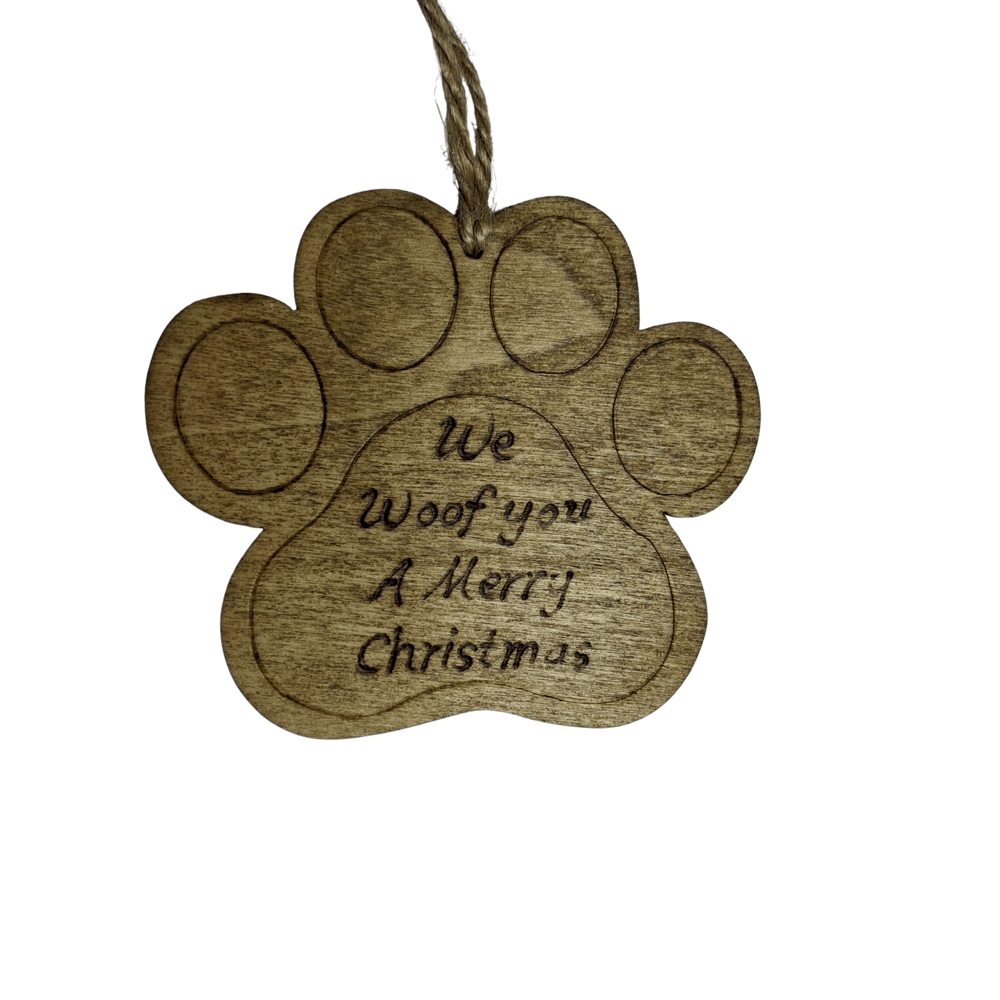A close-up of a brown, wooden ornament shaped like a dog's paw print. It has a hole at the top with a piece of natural twine for hanging. The words "We Woof you A Merry Christmas" are carved into the center of the paw. The background is solid black.