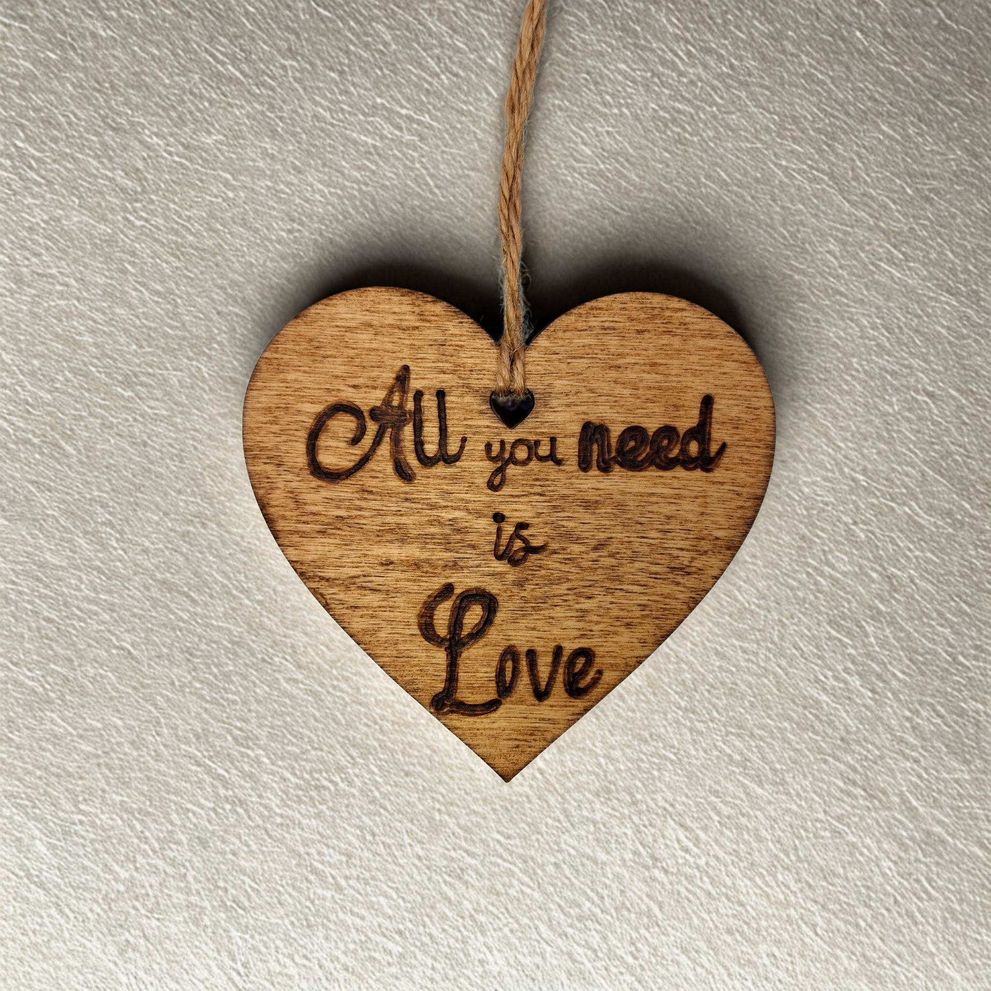 Heart-shaped wooden ornament, hand-drawn with pyrography, featuring the engraved phrase "All you need is Love" in a decorative script, set against a textured background.