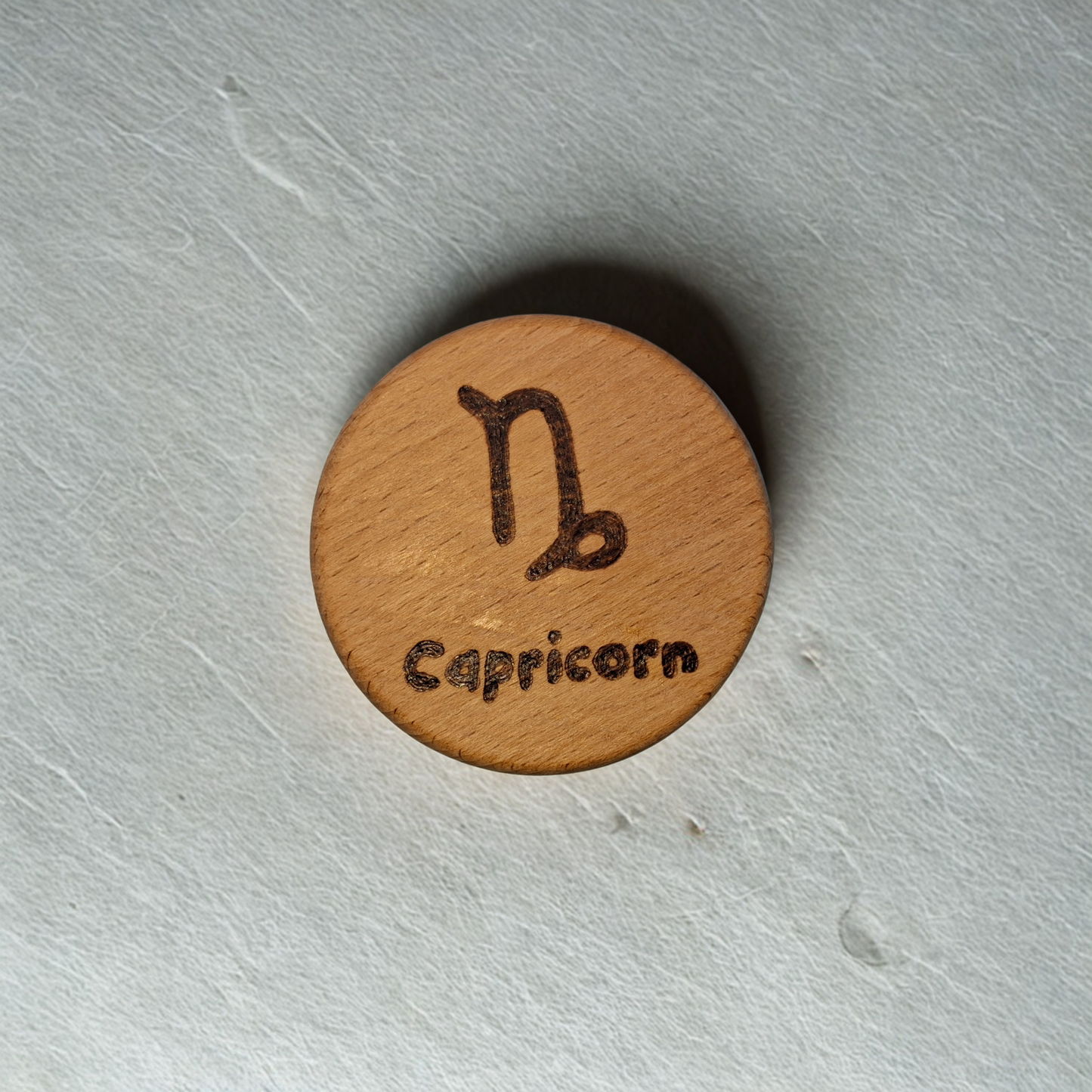 Close-up of a light-colored beechwood circle, 5cm in diameter and 0.4cm thick, featuring the words "Capricorn" and an illustration of the star sign symbol,  hand-burned into its surface using pyrography. The wood has a smooth, waxed finish and the text is clearly legible.