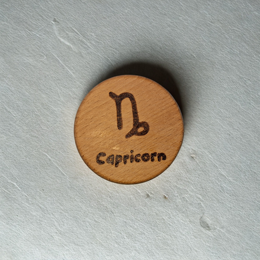 Close-up of a light-colored beechwood circle, 5cm in diameter and 0.4cm thick, featuring the words "Capricorn" and an illustration of the star sign symbol,  hand-burned into its surface using pyrography. The wood has a smooth, waxed finish and the text is clearly legible.
