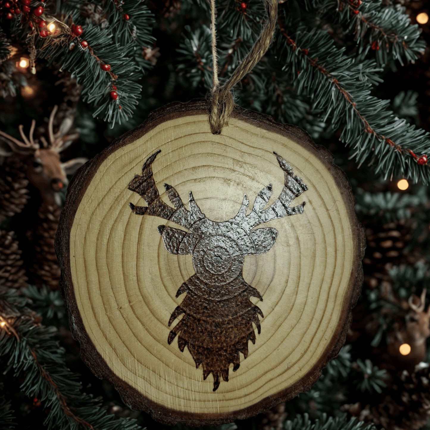 A round Christmas ornament, made from a wooden log slice, is shown on a black background. The ornament is decorated with a pyrography design of a stag's head with large antlers. A jute rope is attached at the top for hanging.
