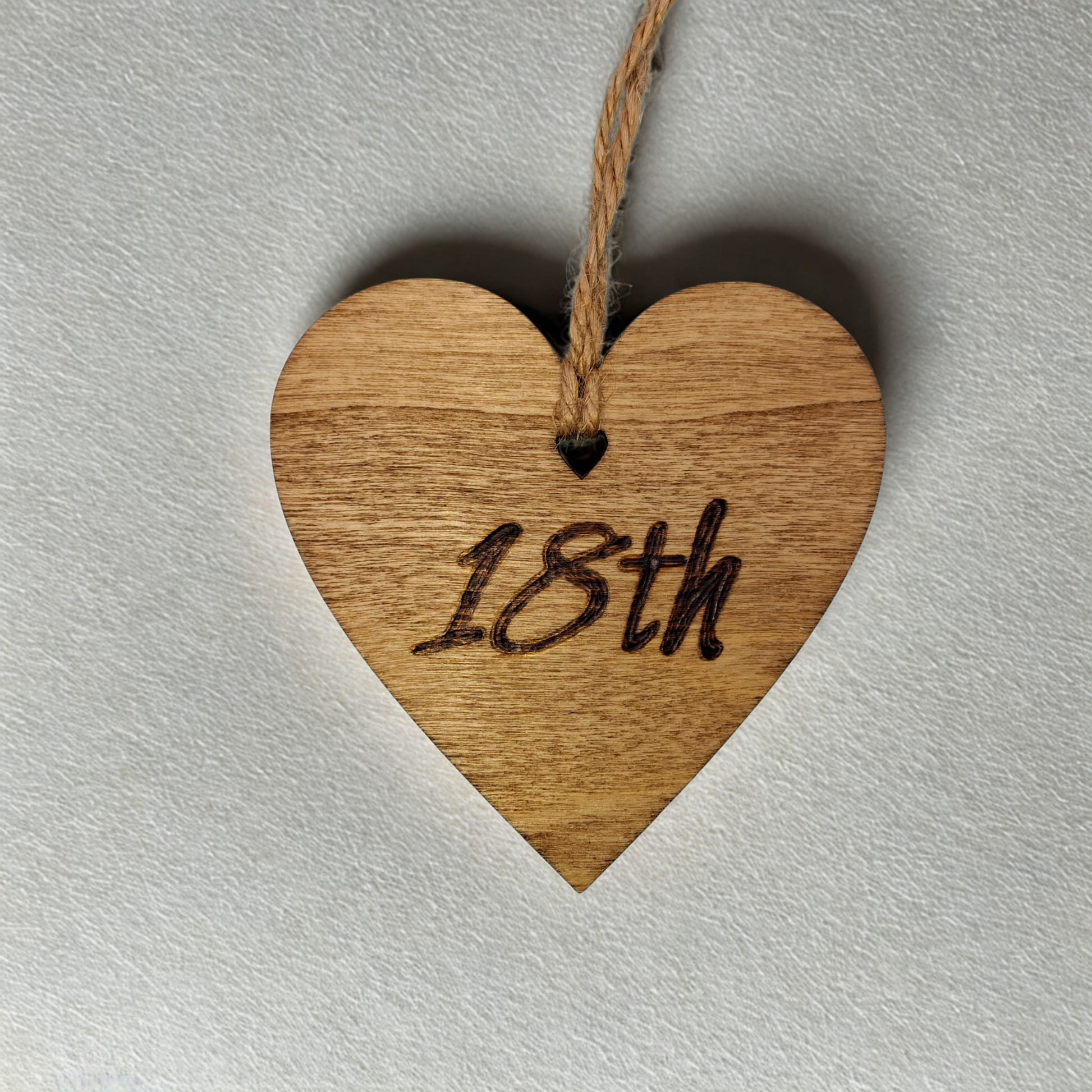 A small,  8cm by 8cm, light-colored wooden heart, decorated with "18th" hand-burned into its surface using pyrography. A short length of natural jute rope is tied through the top for hanging.