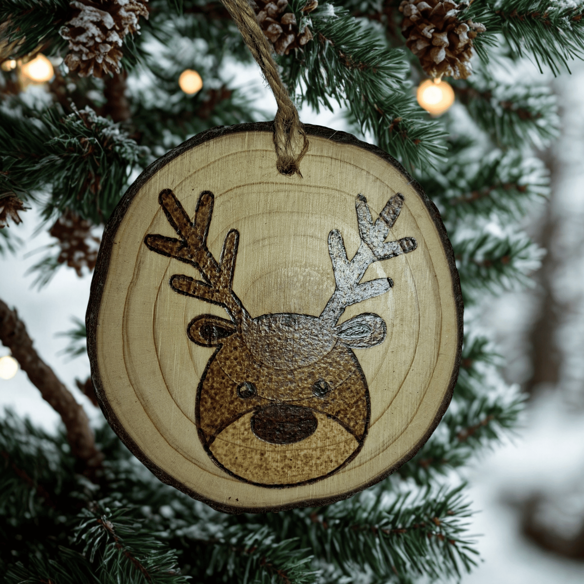 A round Christmas ornament, made from a wooden log slice, is shown on a black background. The ornament is decorated with a pyrography design of a cartoon reindeer's face with antlers. A jute rope is attached at the top for hanging.
