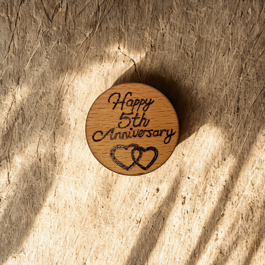 Close-up of a light-colored beechwood circle, 5cm in diameter and 0.4cm thick, featuring the words "Happy 5th Anniversary" and an illustration of 2 hearts hand-burned into its surface using pyrography. The wood has a smooth, waxed finish and the text is clearly legible.