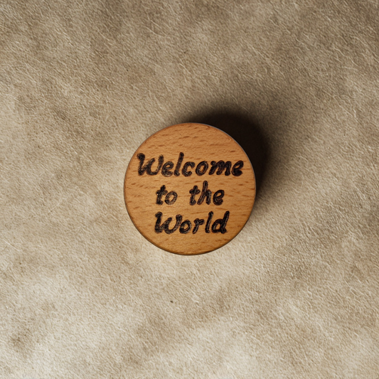 Close-up of a light-colored beechwood circle, 5cm in diameter and 0.4cm thick, featuring the words "Welcome to the world" hand-burned into its surface using pyrography. The wood has a smooth, waxed finish and the text is clearly legible.
