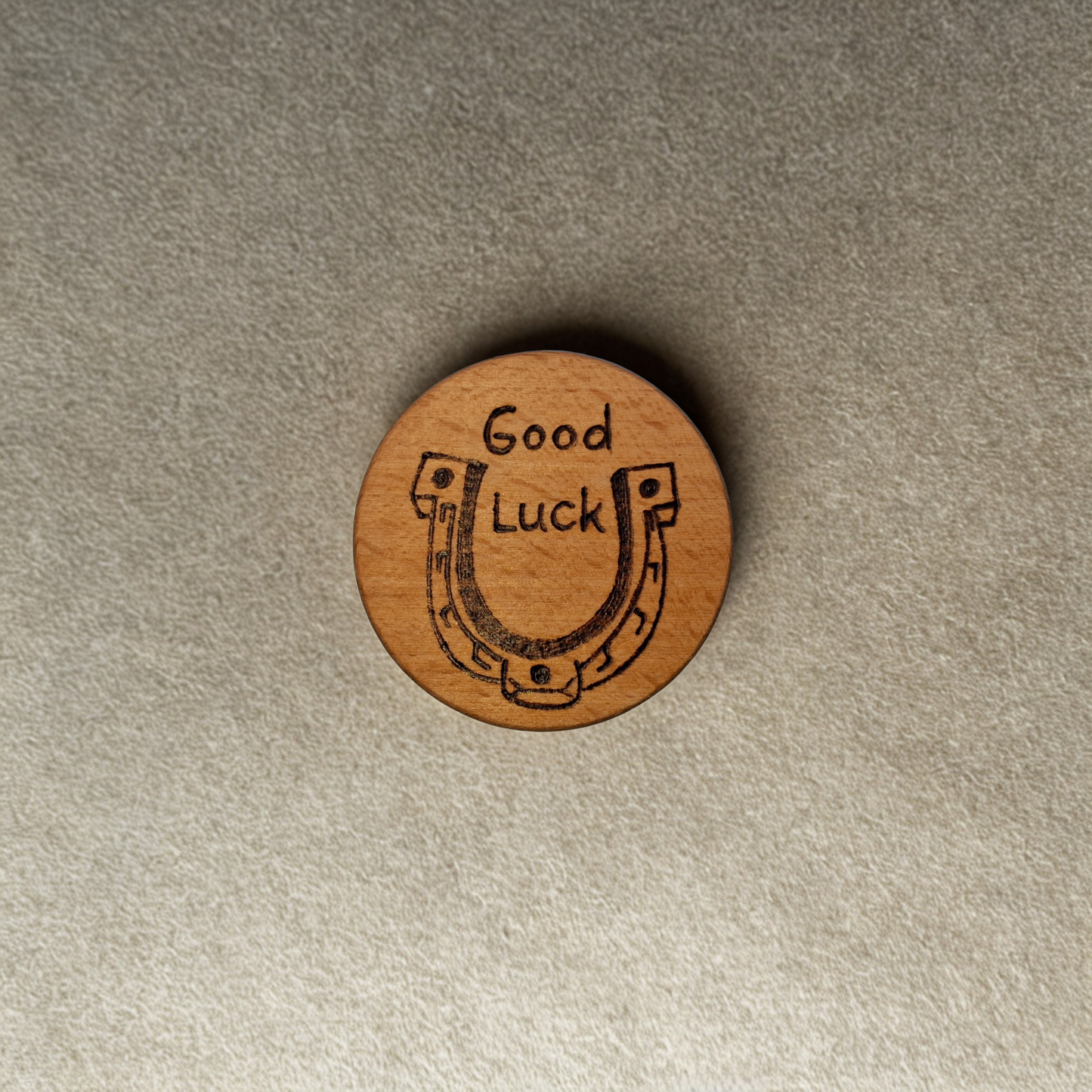 Close-up of a light-colored beechwood circle, 5cm in diameter and 0.4cm thick, featuring the words "Good Luck" and an illustration of a horse shoe hand-burned into its surface using pyrography. The wood has a smooth, waxed finish and the text is clear