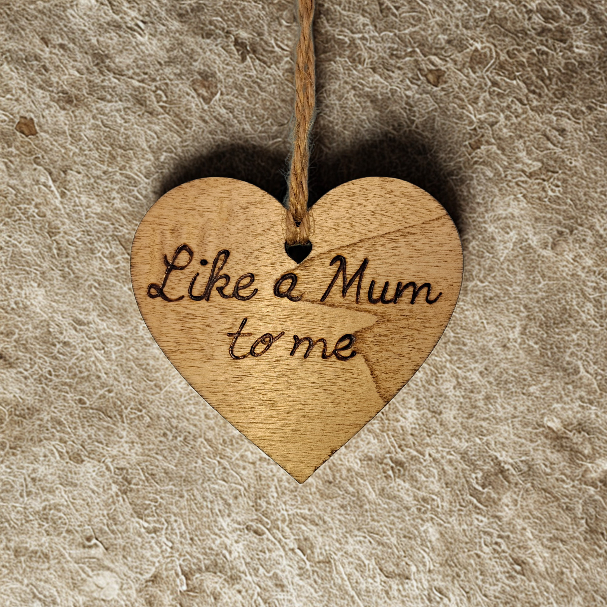 Heart-shaped wooden ornament, hand-drawn with pyrography, featuring the engraved phrase "Like a Mum to me" in a decorative script, set against a textured background.