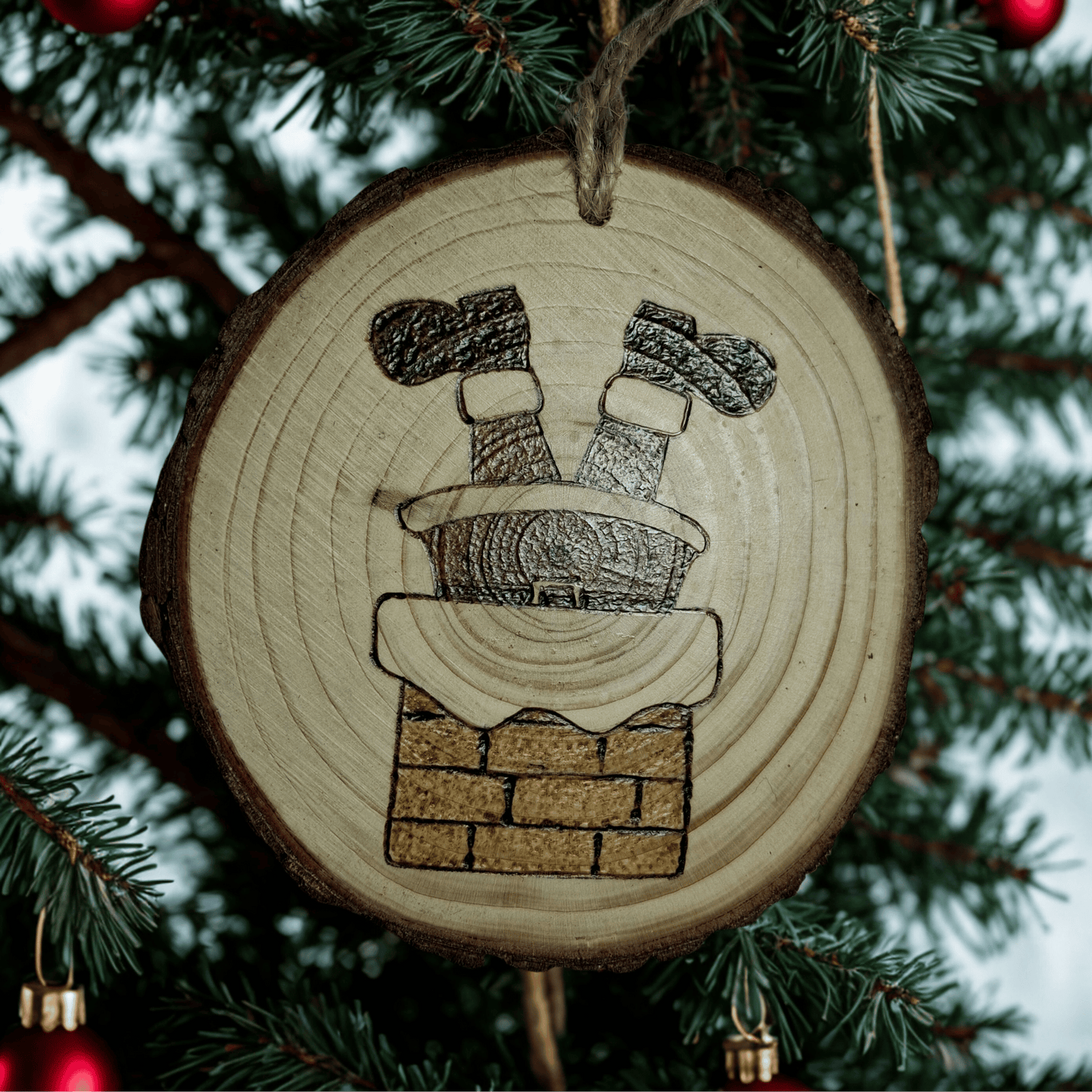 A round wooden Christmas ornament with a jute rope for hanging, centered against a black background. The ornament is decorated with a pyrography design of Santa Claus's legs and boots sticking out of a brick chimney.