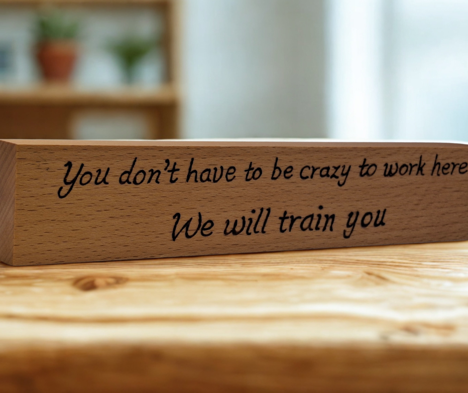 A hand-burnt, freestanding wooden plaque with the words "You don't have to be crazy to work here we will train you" in a casual, handwritten style. The wood has visible grain and a rustic, natural finish.