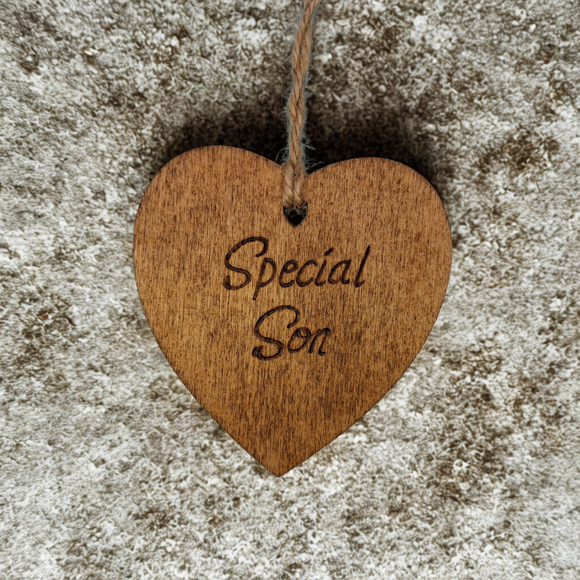 A small, possibly 8cm by 8cm, light-colored wooden heart, decorated with "Special Son" hand-burned into its surface using pyrography. A short length of natural jute rope is tied through the top for hanging.