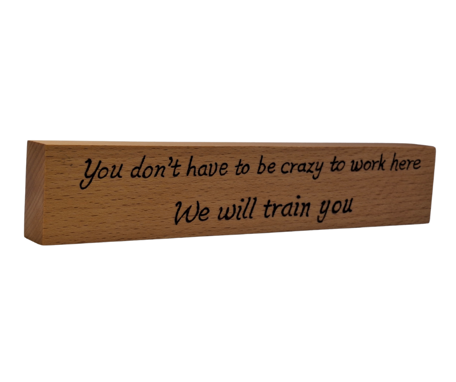 A hand-burnt, freestanding wooden plaque with the words "You don't have to be crazy to work here we will train you" in a casual, handwritten style. The wood has visible grain and a rustic, natural finish.