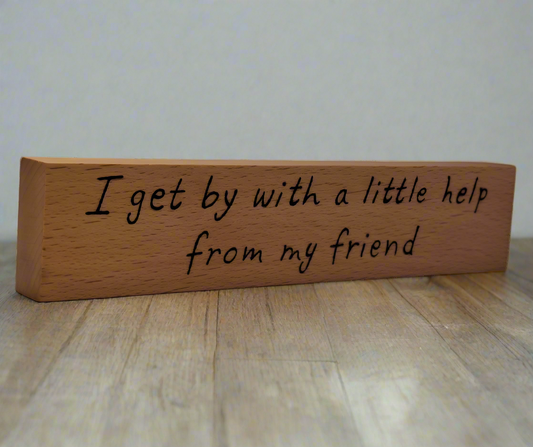 A hand-burnt, freestanding wooden plaque with the words "I get by with a little help from my friend" in a casual, handwritten style. The wood has visible grain and a rustic, natural finish.
